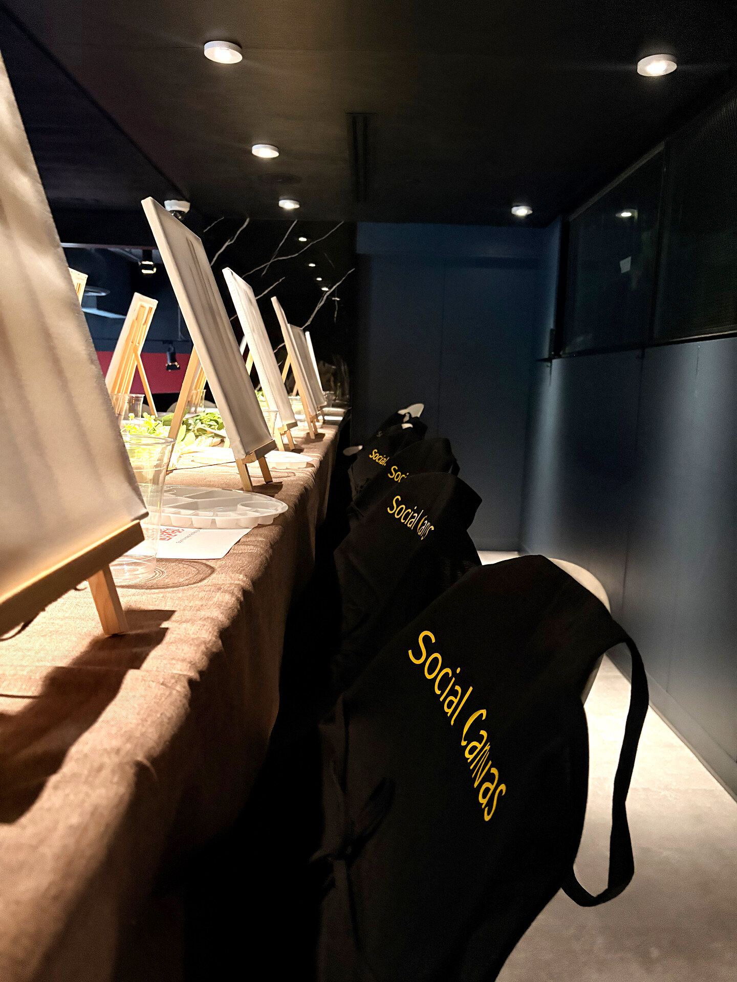Row of easels and branded Social Canvas aprons set up along a long table for a paint and sip night
