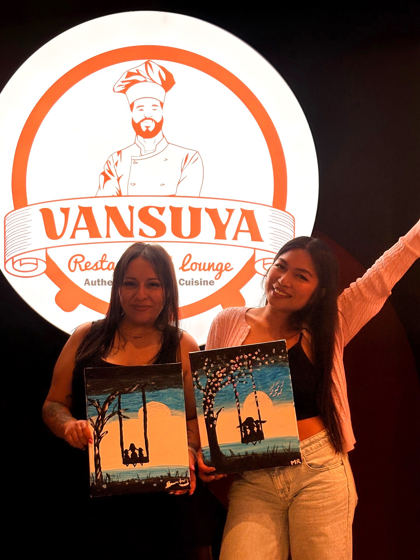 Two guests posing with their sunset swing paintings in front of the VanSuya sign