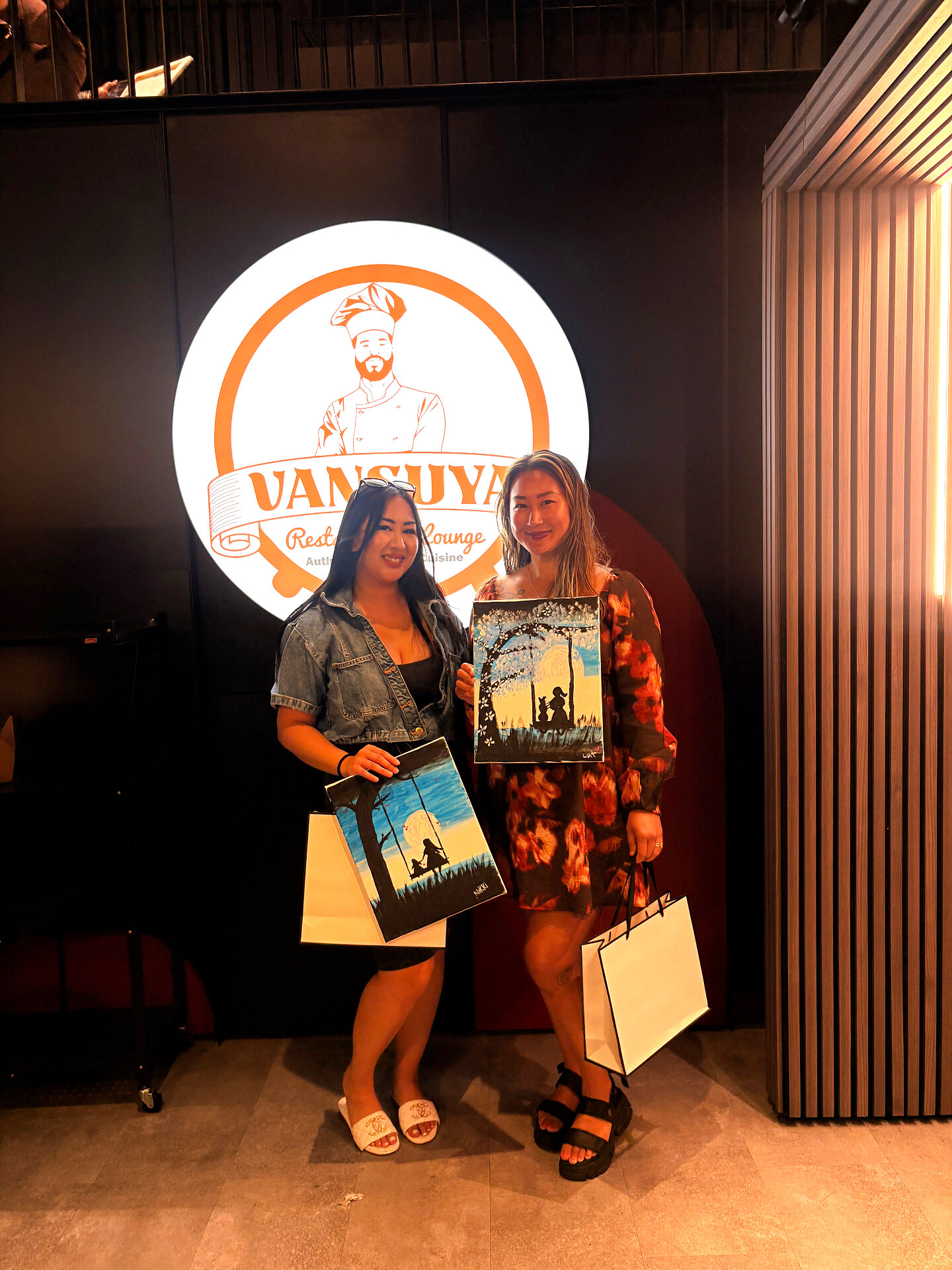 Two friends posing with sunset and moonlit swing paintings in front of the VanSuya logo